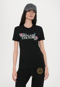 Črna bombažna majica s tiskom "Versace Jeans Couture" obkrožena s cvetličnim vezenjem. V kombinaciji z črnimi trenirkami, ki prikazujejo logotip.