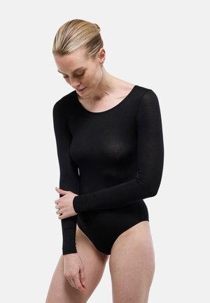 Femme aux cheveux blonds attachés en chignon, portant un body noir à manches longues ajusté, regardant vers le bas et touchant son bras gauche, sur un fond uni.