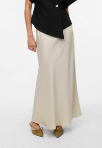 Gonna maxi in satin crema fluido e dalla forma svasata, abbinata a una maglietta nera con un bottone dorato; indossata con scarpe verde oliva decorate con un fiocco.