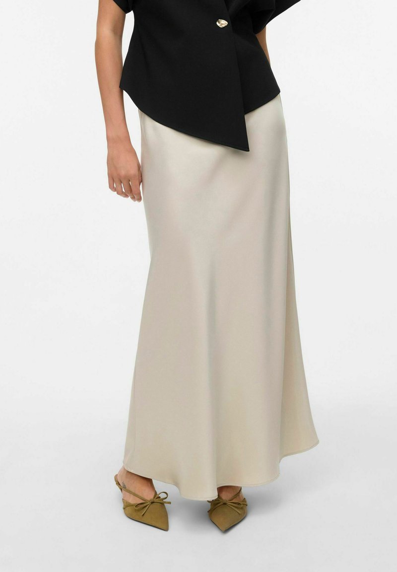 Gonna maxi in satin crema fluido e dalla forma svasata, abbinata a una maglietta nera con un bottone dorato; indossata con scarpe verde oliva decorate con un fiocco.
