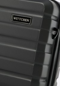 Zwarte harde koffer met een gestructureerd oppervlak, geribbeld ontwerp en een metalen logo plaat. Voorzien van een robuuste rits en zijhandvat.