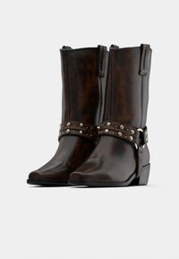 Σκούρα καφέ δερμάτινες μπότες μήκους μέχρι το μέσο της γάμπας με ασ ημένια τρουκς σε ιμάντες γύρω από τους αστραγάλους, χαμηλά τακούνια και θηλιές στο πάνω μέρος.