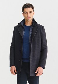 Czarny płaszcz w kratę z wysokim kołnierzem, z zapięciem na guziki. Pod nim widać granatowy sweter. Gładka tekstura, dopasowany krój.