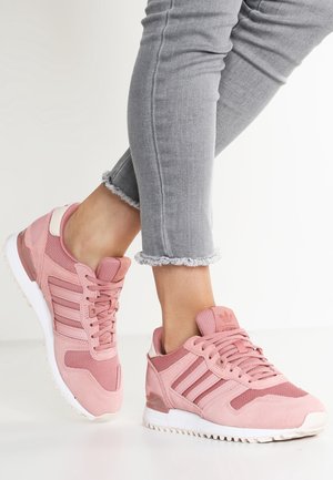 Osoba nosząca jasnoszare dżinsy z postrzępionym dołem i różowe buty sportowe Adidas z białą podeszwą, stojąca na białej podłodze.