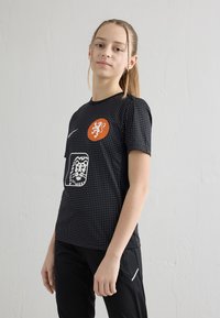 Adolescente com cabelo trançado, vestindo camisola e calças desportivas pretas com bolinhas, em pé contra uma parede branca texturizada, olhando para a câmara.