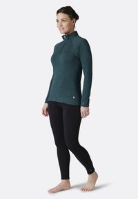 Teal long-sleeve top with a half-zip collar, paired with black fitted leggings. Top features subtle texture and logo detail on the hem.