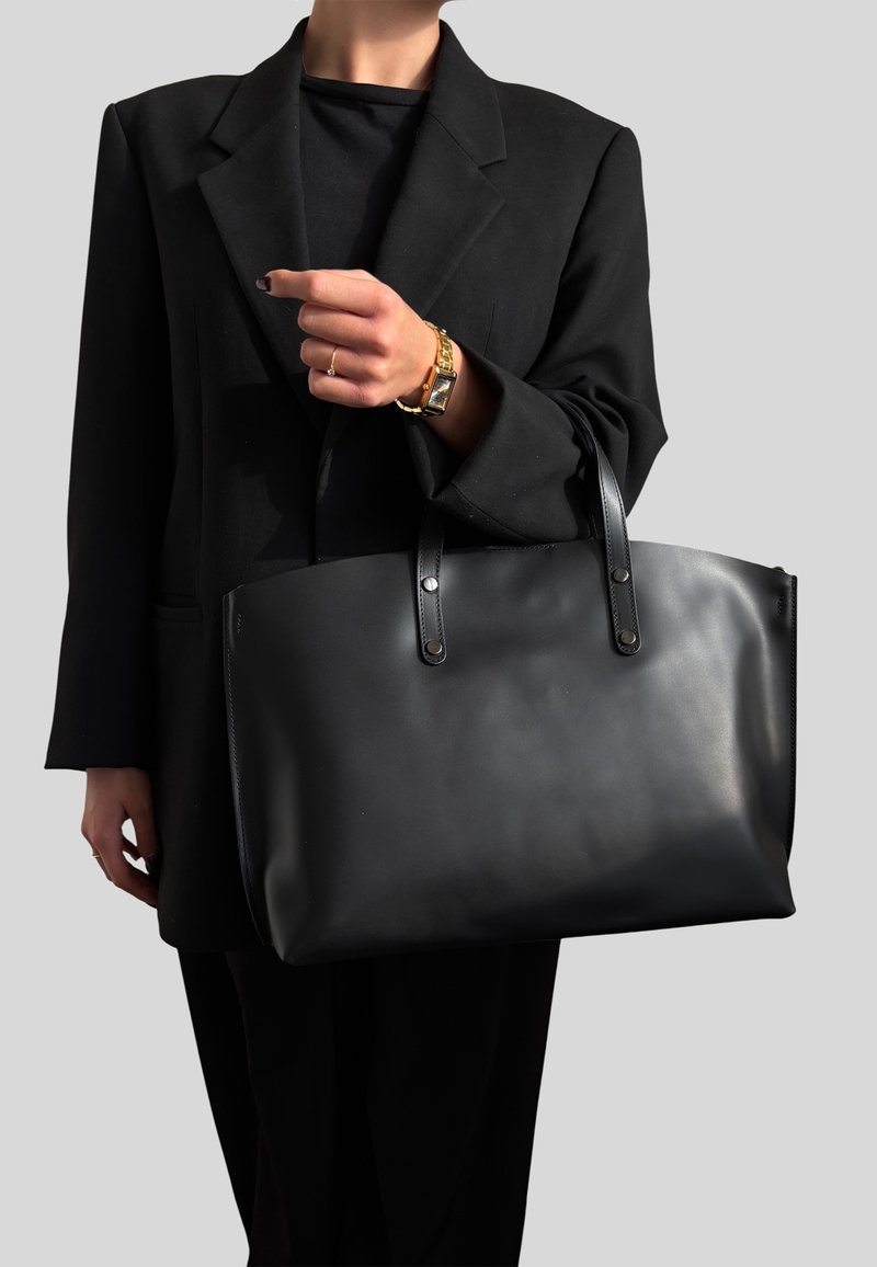 Personne portant un blazer et un pantalon noirs tient un grand sac cabas en cuir noir avec des poignées sur fond gris.