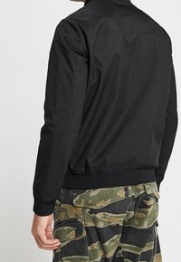 Svart bomberjacka med en slät yta, ribbade ärmslut och midja. Har en enkel, klassisk design utan ytterligare dekorationer.