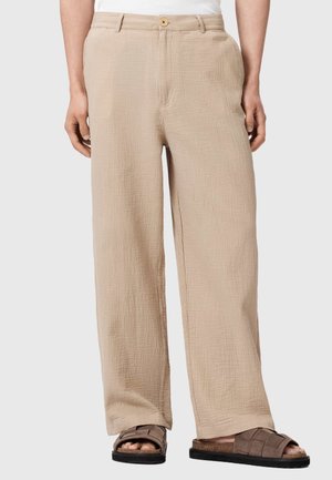 Beżowe, teksturowane spodnie z szerokimi nogawkami i guzikami z przodu, zestawione z brązowymi plecionymi sandałami wsuwanymi na stojącej osobie.