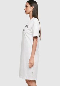 Rochie din bumbac alb, cu mâneci scurte, guler rotund și un logo imprimat pe față. Dispune de un buzunar lateral cu un accent verde.