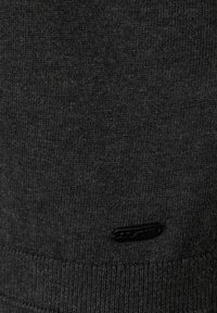 Mörkgrått stickat tyg med ribbat mönster, med en liten svart logotypetikett sydd vid nederkanten. Enkel och funktionell design.