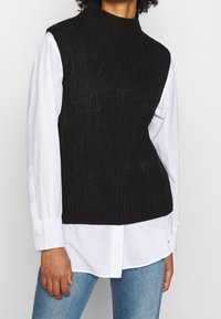 Schwarze gestrickte ärmellose Rollkragenweste, geschichtet über einem weißen Langarmshirt. Verfügt über eine gerippte Textur und eine lockere Passform.