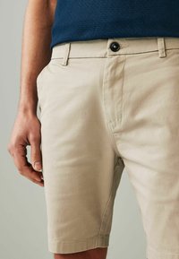 Beige chinos egyenes szabással, elülső gombos zipzárral, oldalsó zsebekkel és sima pamutszövetből, csípőszinten bemutatva.