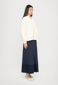 Svetlá béžová mikina na zips s kapucňou a oversized strihom. Tmavomodrá maxi sukňa s kontrastným lemom. Model predvádzajúci smotanové muly.