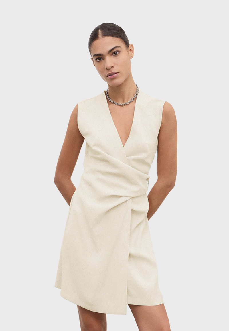 Femme portant une robe beige sans manches à col en V profond et un collier en chaîne argentée, debout les mains derrière le dos.