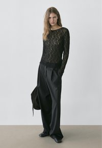 Black lace long-sleeve top with a textured pattern; paired with loose-fitting black trousers and slip-on shoes. Minimalist design, neutral colors.