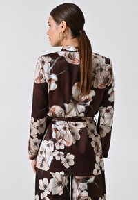 Bloemmotief, zijden bruine blouse met lange mouwen en een ceintuur in de taille. Heeft lichtgekleurde bloemen en een gladde textuur.