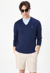 Navy v-neck sweater in a soft knit material, paired with beige pinstriped shorts. Model wearing dark sunglasses, hands in pockets.