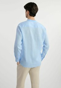 Jasnoniebieska koszula z długim rękawem wykonana z lnu, z subtelną fakturą, zaokrąglonym dołem i mankietami na guziki. W zestawie z beżowymi spodniami.