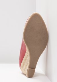 Semelle d'une chaussure compensée rouge taille 37 avec une semelle intermédiaire en jute tressé et une semelle extérieure en caoutchouc texturé, inclinée contre un coin blanc.