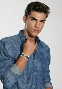 Blaue Jeanshemd mit einem strukturierten Muster, hochgekrempelten Ärmeln und metallischen Akzenten. Trägt mehrere farbige Armbänder am Handgelenk.