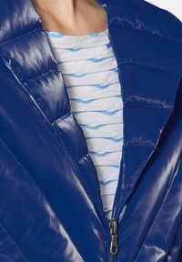 Glänzende blaue Pufferjacke mit Reißverschluss und strukturiertem Kragen. Darunter ist ein hellblaues Oberteil mit weißem Muster sichtbar.