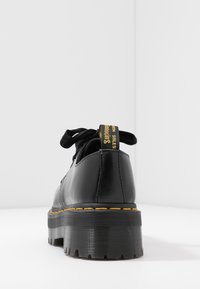 Scarpe in pelle nera con suola spessa e scanalata, cuciture gialle e design con lacci neri. Presentano un plug con testo giallo.