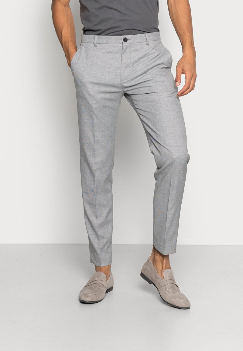 Uomo che indossa pantaloni eleganti grigi slim-fit con tasche frontali, abbinati a mocassini in suede grigio chiaro, in piedi di fronte a uno sfondo semplice.
