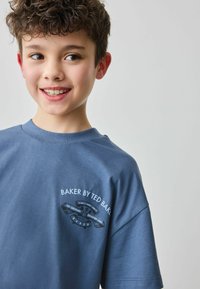 Zils kokvilnas T-krekls ar apaļu kakla izgriezumu un īsām piedurknēm. Ievērojams grafiskais izdrukas attēls uz krūšu daļas ar tekstu "BAKER BY TED BAKER."