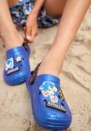 Enfant assis sur le sable portant des sabots bleus décorés avec Sonic the Hedgehog et d'autres breloques sur le thème des dessins animés.