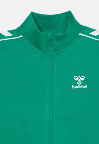 Roheline lukuga sportjakk, millel on vasakul rinnal valge hummeli logo ja valged ševroontriibud õlgadel.