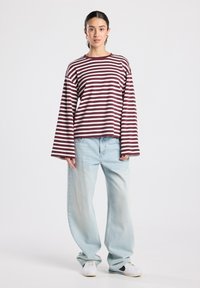 Langärmliges gestreiftes Oberteil in Burgunderrot und Weiß, kombiniert mit hellblauen Jeans mit weitem Bein. Glatter Stoff mit entspanntem Schnitt und überschnittener Schulter.