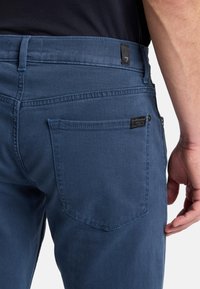 Jeans in denim blu da uomo con una trama liscia, taglio dritto, cuciture visibili e un'etichetta a toppa sulla tasca posteriore.