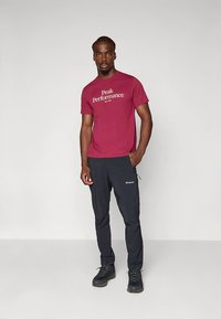 Vínovo červené tričko s logom "Peak Performance", čierne nohavice a čierne turistické topánky. Oblečenie je praktické a určené na vonkajšie aktivity.