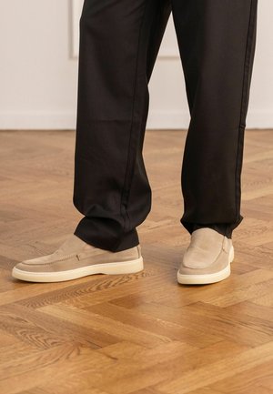 Persoon draagt beige suède instapschoenen met witte zolen en loszittende zwarte broek, staand op een houten parketvloer.