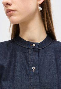 Γυναίκα που φοράει μπλε σκουρόχρωμο πουκάμισο με κουμπιά και γιακά μπαντάνα, καθώς και ένα σκουλαρίκι σε σχήμα ασημένιας σταγόνας στο αριστερό της αυτί.