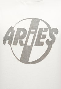 Γκρι κείμενο Κριός με διαγώνια ρίγα μέσα σε κύκλο σε λευκό ύφασμα, που μοιάζει με επώνυμο λογότυπο ή σχέδιο σε ρούχα.