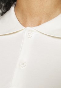 White polo shirt featuring a textured fabric, a classic collar, and two matching buttons at the neckline.