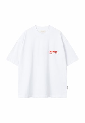 Camiseta blanca de manga corta con texto rojo "another." en la parte superior izquierda del pecho y una pequeña etiqueta de tela en la costura lateral izquierda.