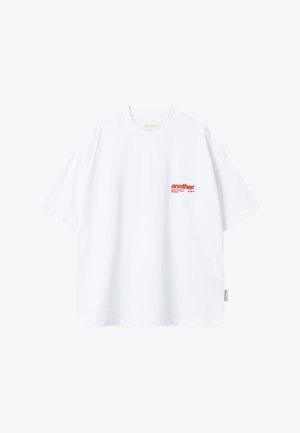 Camiseta blanca de manga corta con texto rojo "another." en la parte superior izquierda del pecho y una pequeña etiqueta de tela en la costura lateral izquierda.