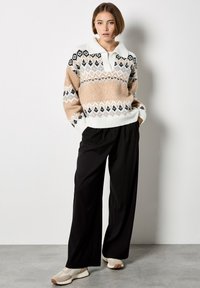 Sweter z dzianiny z wzorzystym deseniem w odcieniach beżu, czerni i bieli, z kołnierzem. W komplecie z czarnymi szerokimi spodniami i jasnymi trampkami.