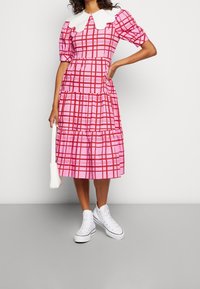 Femme portant une robe midi à carreaux rose et rouge avec un col Peter Pan blanc, des baskets blanches montantes et portant un sac à main blanc.