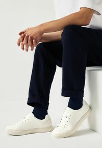 White textured sneakers with flat soles, paired with rolled navy pants and dark socks. The setting has a neutral background.