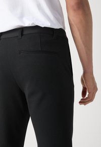 Black tailored trousers with a smooth texture, featuring a back pocket and side pockets, paired with a white shirt.