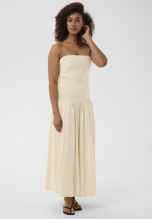 Femme aux cheveux bouclés portant une longue robe crème sans bretelles et des sandales noires, debout devant un fond blanc uni.