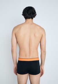 Caleçons boxers noirs avec une taille orange, présentant un tissu lisse et un design ajusté, vus de dos. Peau visible au-dessus de la taille.