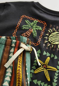 Close-up van zwarte stof met geborduurde palmboom, zon en zeester, naast gestreepte stof met een crèmekleurig trekkoord.