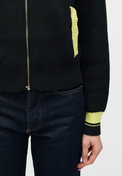 Μαύρο ριμπ πουλόβερ με φερμουάρ, με φωτεινές κίτρινες λεπτομέρειες στην τσέπη και τα μανίκια, φορεμένο με σκούρο μπλε τζιν, με το χέρι στο πλάι.