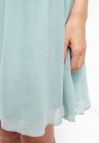 Světle mátově zelené šaty vyrobené z průsvitné látky, s volným střihem a mírně zvlněným lemem, doplněné jemnou texturou.