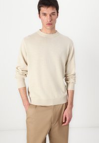 Μπεζ πλεκτό πουλόβερ με στρογγυλή λαιμόκοψη, ραμμένα μανίκια και τελείωμα, φορεμένο πάνω από ανοιχτό καφέ παντελόνι. Μαλακή υφή και άνετη εφαρμογή.